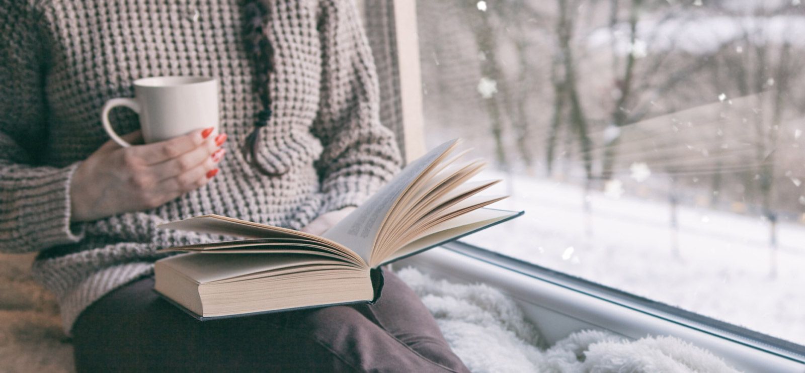 Dame sitter i vinduskarmen med en hvit kopp og en bok.